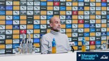 A person is seated at a press conference with a Manchester City sponsor backdrop. Bottles of water and a Lucozade drink are on the table.