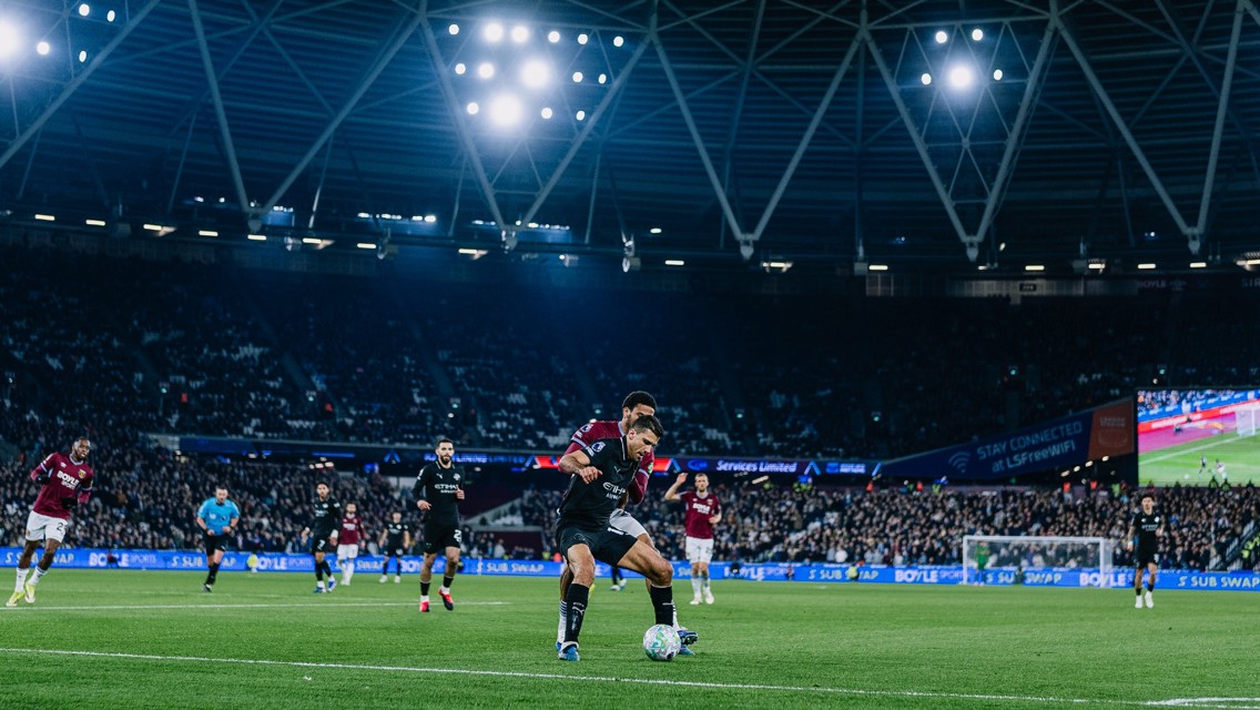 A night stadium soccer match with players in action, one controlling the ball near the sideline. The stadium is brightly lit, filled with spectators.
