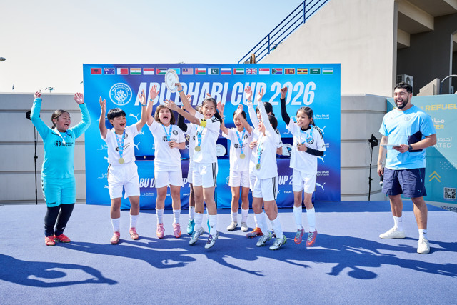 Young athletes celebrating at the Abu Dhabi Cup 2026 award ceremony, organized by Manchester City, wearing white team uniforms with medals. Flags from different countries are displayed in the background.