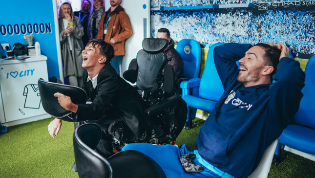 Group of people in a room decorated with Manchester City theme. Two people are playing video games. One is positioned in a wheelchair. Gaming controllers are visible on their laps.