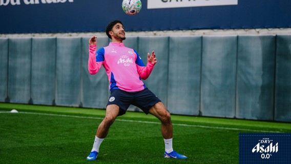 A soccer player in a pink training kit maneuvering to control the ball in mid-air on a grassy field. Advertising boards are visible in the background.