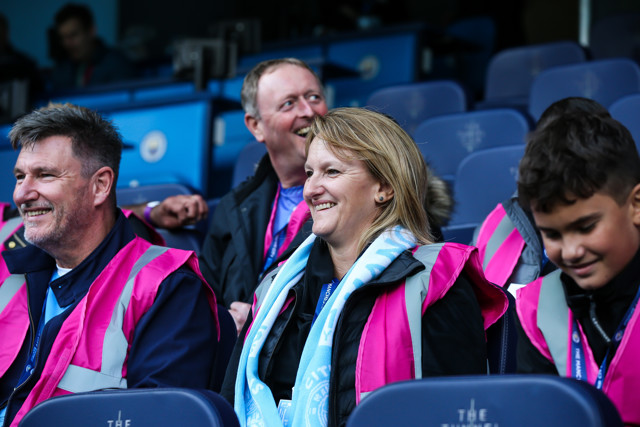 Four individuals wearing bright pink vests sitting in stadium seats with Manchester City branding.