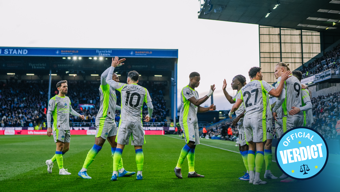 A soccer team wearing gray and neon uniforms huddled on the field celebrating with high-fives in front of a filled stadium stand. There is a circular badge with 'Official Verdict' in the bottom right corner.