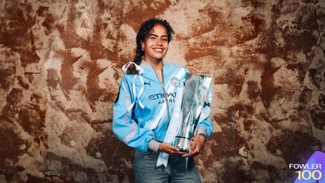 A woman wearing a Manchester City football jersey holding a silver Barclays trophy against a textured brown background.