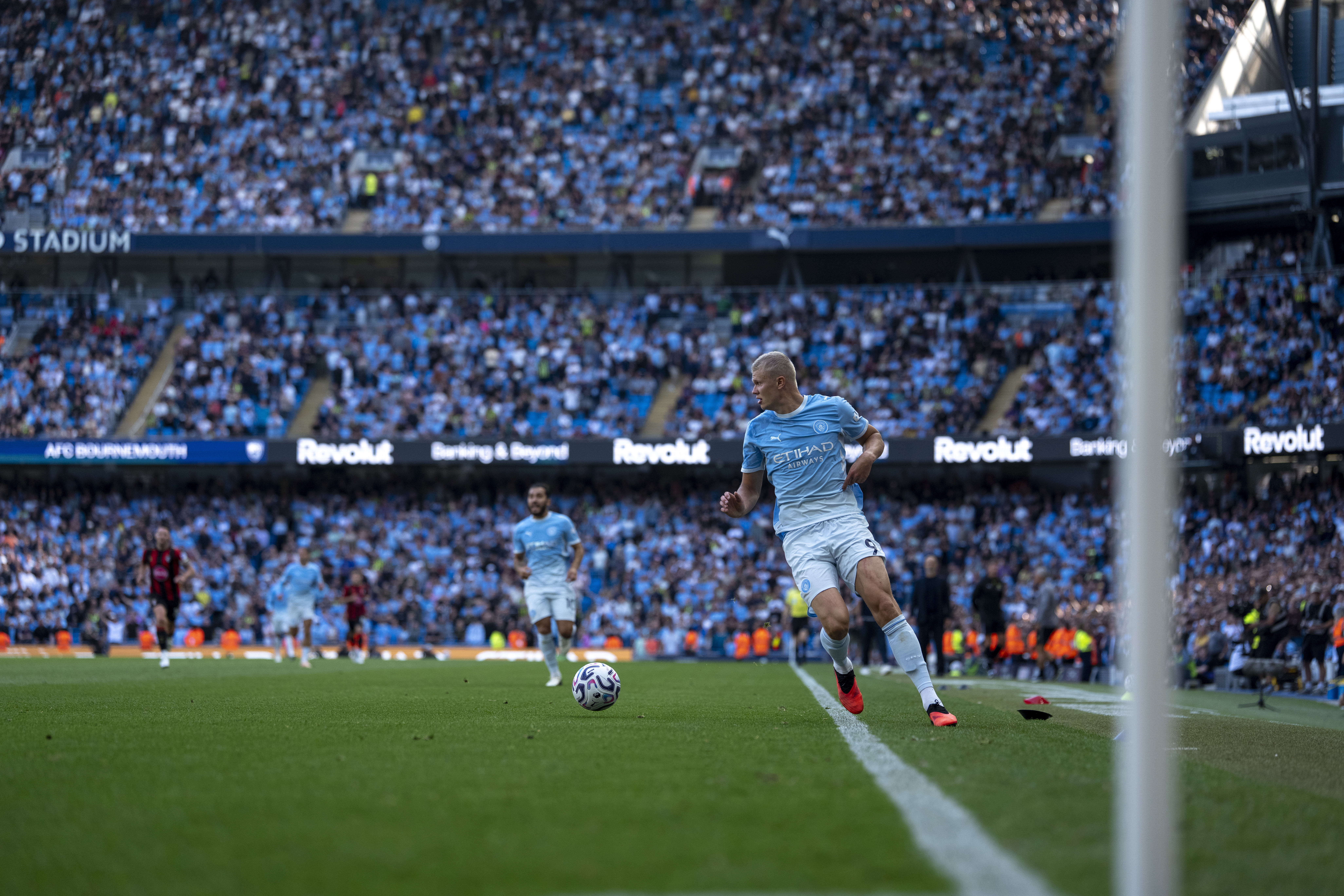 A Manchester City player is dribbling the ball during a match at Etihad Stadium, with a large crowd in the background.