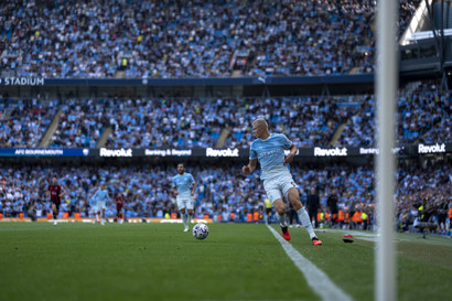 A Manchester City player is dribbling the ball during a match at Etihad Stadium, with a large crowd in the background.