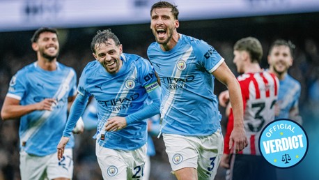 Manchester City players in blue jerseys celebrating during a match against a team in red and white kits. One player has the captain's armband.