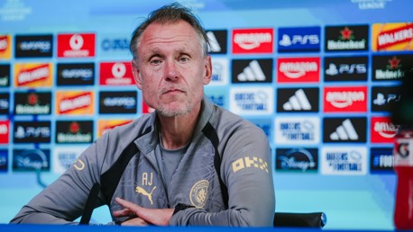 A person in a Manchester City press conference setting, wearing a jacket with a club logo and sponsor logos in the background.
