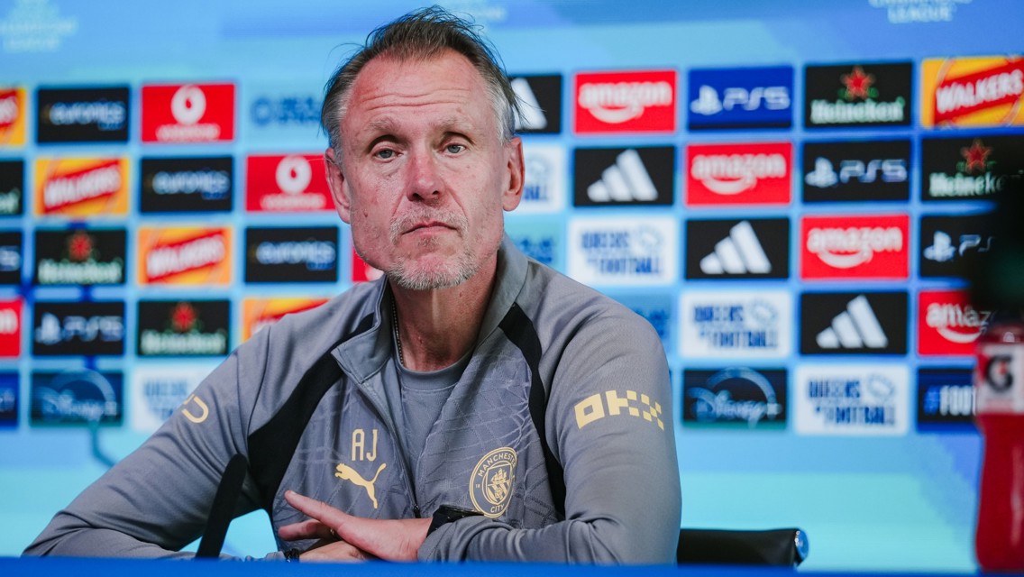 A person in a Manchester City press conference setting, wearing a jacket with a club logo and sponsor logos in the background.