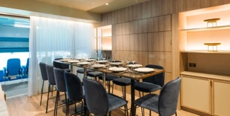 An elegant dining room with a long table set for dining, featuring blue cushioned chairs and wooden paneling on the walls. Shelves with soft lighting decorate the space.