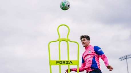 A soccer player in a pink and blue training kit practices with a ball and a neon green training mannequin labeled 'FORZA'. The player is heading the ball during practice on an outdoor field.