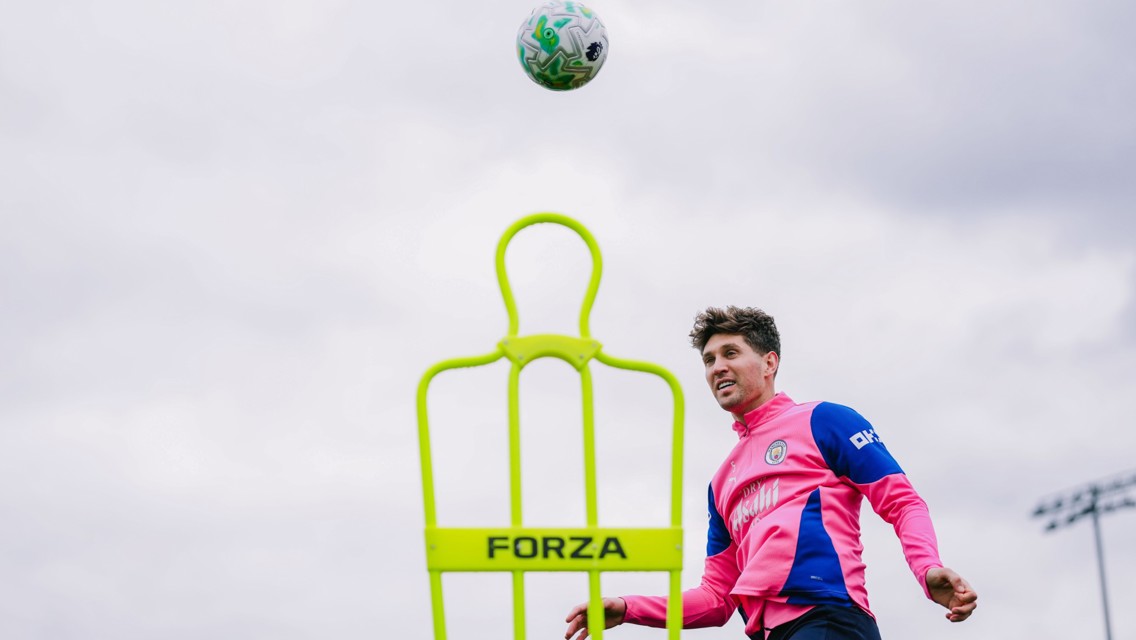 A soccer player in a pink and blue training kit practices with a ball and a neon green training mannequin labeled 'FORZA'. The player is heading the ball during practice on an outdoor field.