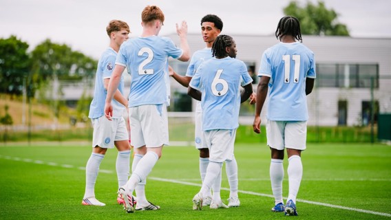 Five soccer players in light blue jerseys and white shorts gather on a field. Two players, numbered 2 and 6, are interacting. The setting suggests a team practice or match preparation.