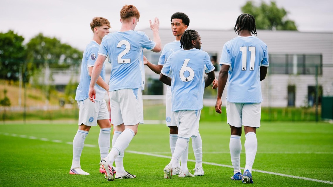Five soccer players in light blue jerseys and white shorts gather on a field. Two players, numbered 2 and 6, are interacting. The setting suggests a team practice or match preparation.