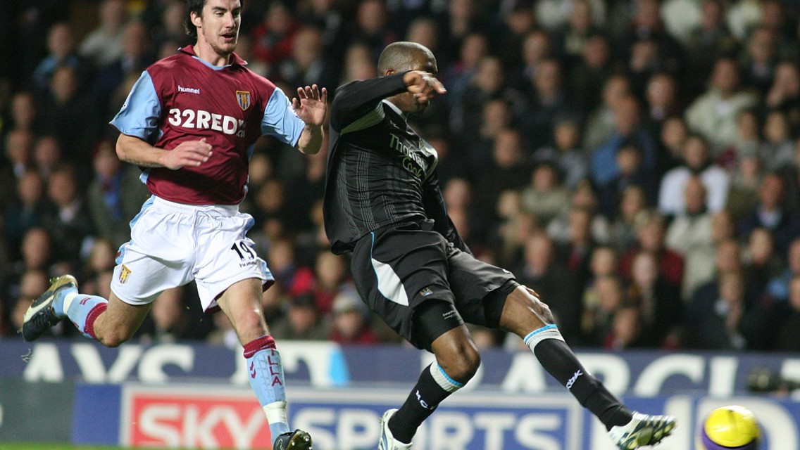 A football match scene with a player from Aston Villa in maroon kit playing against a player in a black kit. The player in black is kicking the ball while the Aston Villa player is defending.
