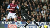 A football match scene with a player from Aston Villa in maroon kit playing against a player in a black kit. The player in black is kicking the ball while the Aston Villa player is defending.