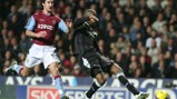 A football match scene with a player from Aston Villa in maroon kit playing against a player in a black kit. The player in black is kicking the ball while the Aston Villa player is defending.