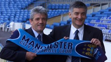 Two men holding a Manchester City Football Club scarf in a stadium. They are wearing suits and ties.