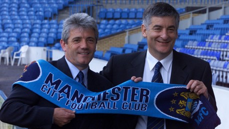 Two men holding a Manchester City Football Club scarf in a stadium. They are wearing suits and ties.