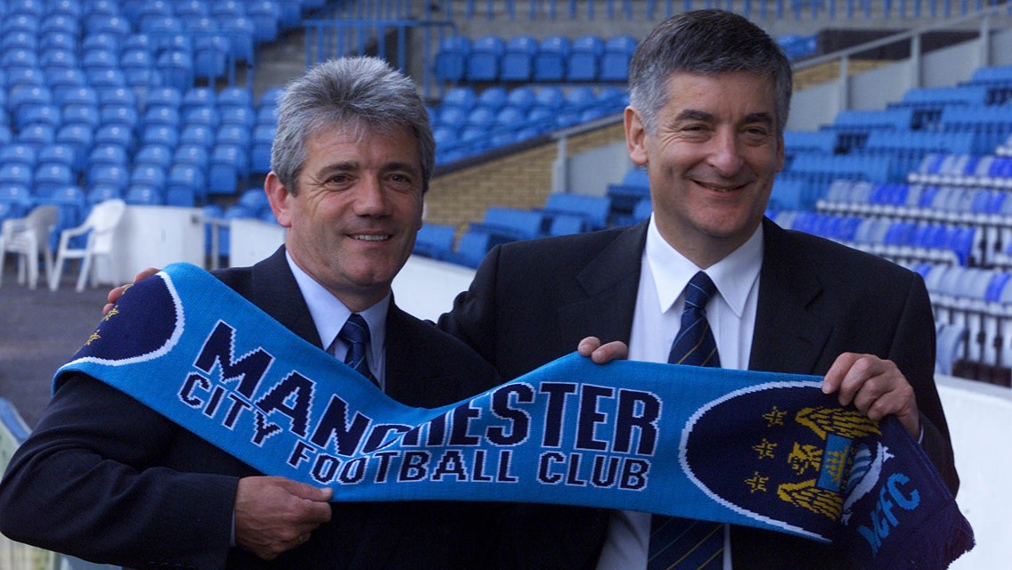 Two men holding a Manchester City Football Club scarf in a stadium. They are wearing suits and ties.