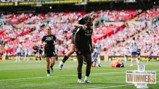 Football players celebrate on the field with one player giving another a piggyback. The background shows a stadium with a crowd. A graphic reads 'Adobe Women's FA Cup Winners 2026'.