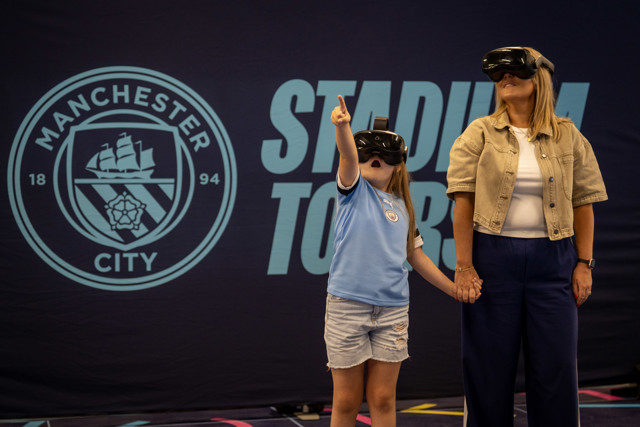 A child and an adult participate in a VR experience at a Manchester City stadium tour, with both wearing VR headsets and standing near a Manchester City logo backdrop.