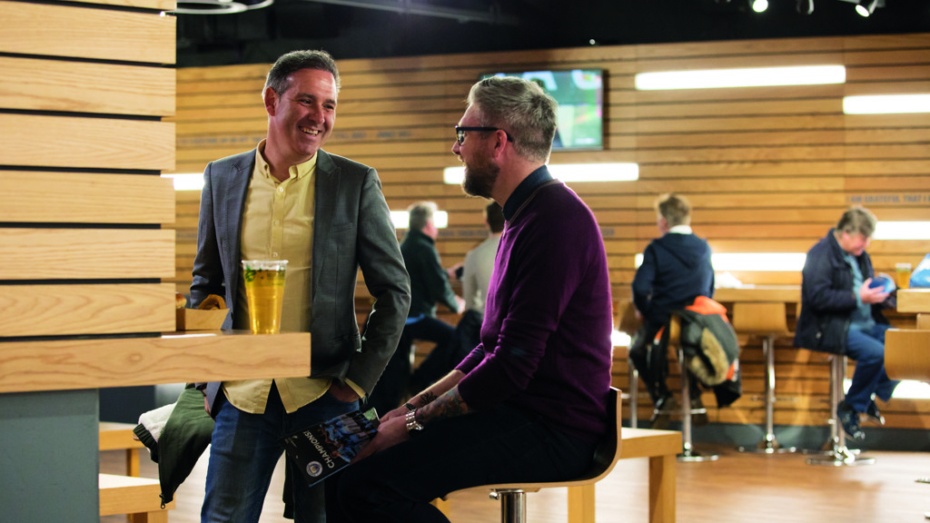 Two men in casual attire having a friendly conversation in a modern lounge area with wood paneling and stools.