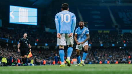 Soccer players in Manchester City kits celebrate a goal in a stadium with a score display in the background.