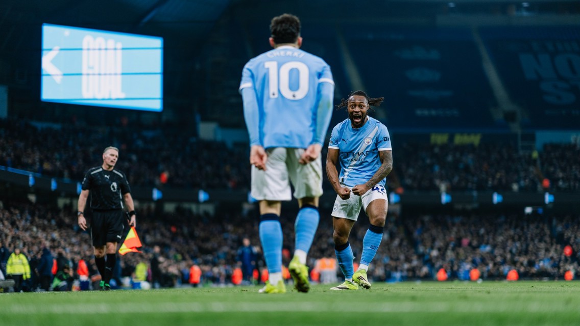Soccer players in Manchester City kits celebrate a goal in a stadium with a score display in the background.
