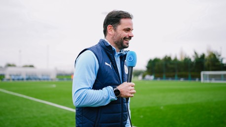 A man with blurred face holding a microphone on a soccer field. He's wearing a blue sleeveless vest over a light blue long-sleeve shirt. The microphone has a Manchester City logo.