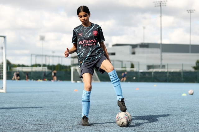 A young football player wearing a Manchester City 2023/24 away kit is on a training ground, balancing a soccer ball with one foot while giving a thumbs-up gesture. The training ground features blue artificial turf with several players and goal posts in the background.