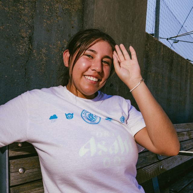 A person smiling and saluting, wearing a white Manchester City t-shirt while sitting on a bench.