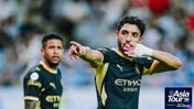 Two football players in black Manchester City kits on a field, one pointing with a gloved hand, the other looking on. Branding for Asia Tour '26 visible.