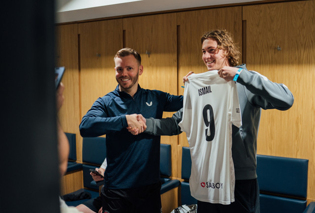 Two players shaking hands in a locker room, with one player holding up a jersey with the name 'Ismail' and the number 9.