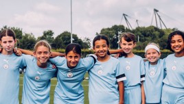 A group of seven children wearing Manchester City football kits standing together on a field with trees and a structure in the background.