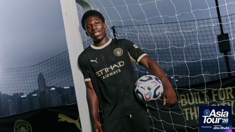 A Manchester City player in black kit holding a soccer ball on an outdoor field with a net and skyline in the background. The image includes Asia Tour '26 branding.