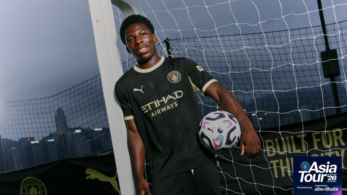 A Manchester City player in black kit holding a soccer ball on an outdoor field with a net and skyline in the background. The image includes Asia Tour '26 branding.