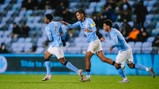 Three Manchester City youth players wearing light blue jerseys celebrate a goal on the field during a match with blurred faces.