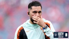 A football player in a jacket with a hand covering mouth, wearing a watch and earbuds. The image has a 'City at the World Cup' logo.