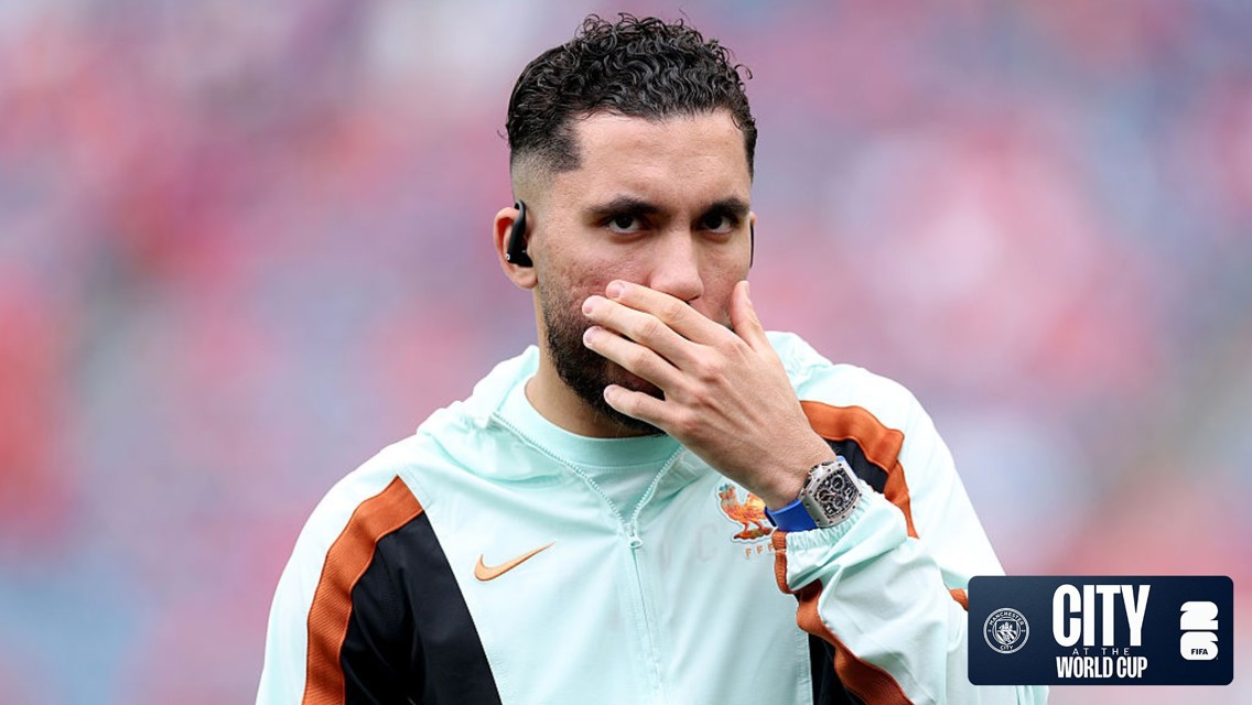 A football player in a jacket with a hand covering mouth, wearing a watch and earbuds. The image has a 'City at the World Cup' logo.