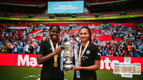 Two players in Manchester City jerseys hold the FA Cup trophy celebrating their win in 2026. They are wearing medals, with the crowd in the background inside a stadium.