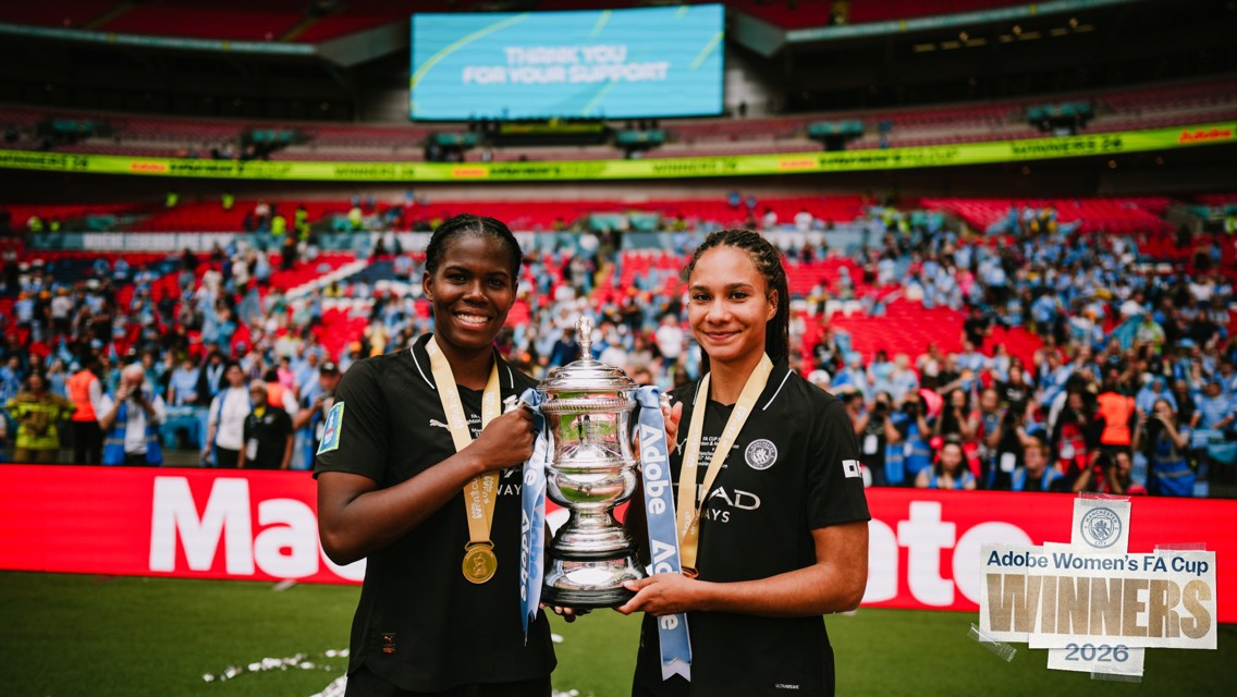 Two players in Manchester City jerseys hold the FA Cup trophy celebrating their win in 2026. They are wearing medals, with the crowd in the background inside a stadium.