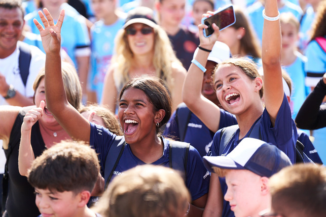 A group of people wearing blue t-shirts, some raising their hands, participating in an outdoor event.
