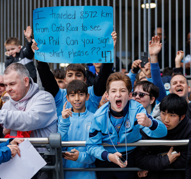 A Manchester City fan holding a sign that reads 'I traveled 8,572 km from Costa Rica to see you Phil. Can you sign my shirt please?' in a crowd of supporters.