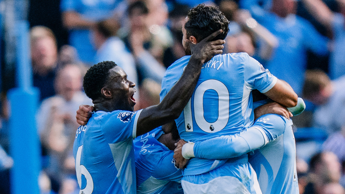 Manchester City players celebrating a goal together during a match, wearing blue jerseys. The player with jersey number 10 is visible.