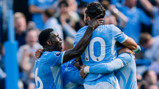 Manchester City players celebrating a goal together during a match, wearing blue jerseys. The player with jersey number 10 is visible.