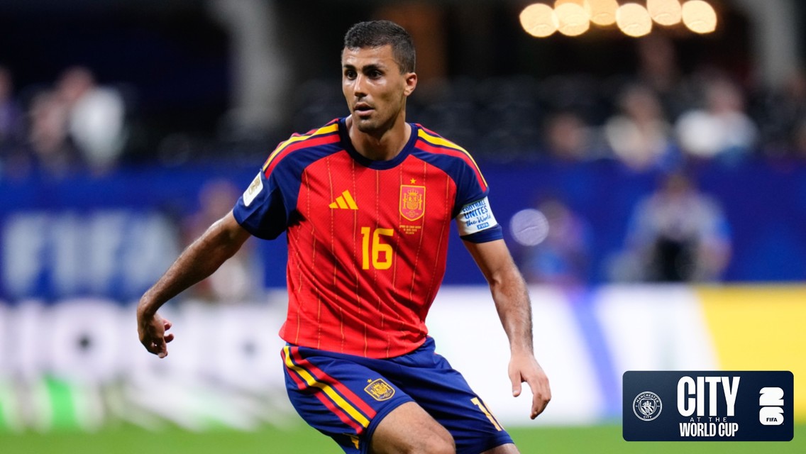 A football player wearing a red and blue Spanish jersey with the number 16 is in action during a World Cup match. The stadium background features blurry spectators.