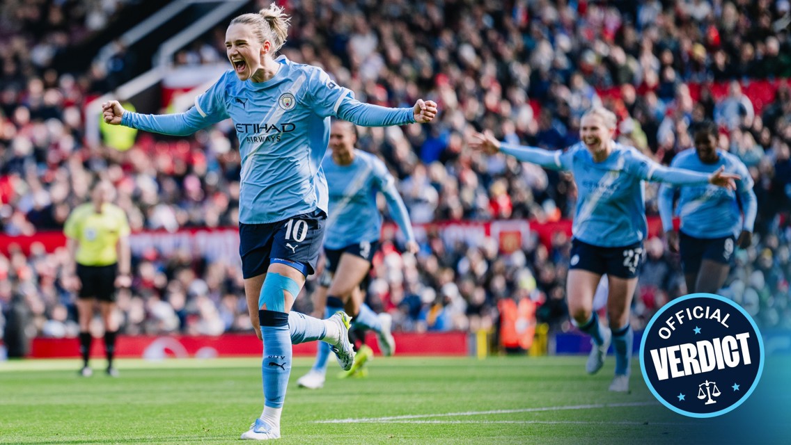 Soccer players wearing Manchester City kits celebrating on the field in front of a cheering crowd. A player wearing number 10 from Manchester City is the focus, with others blurred in the background.