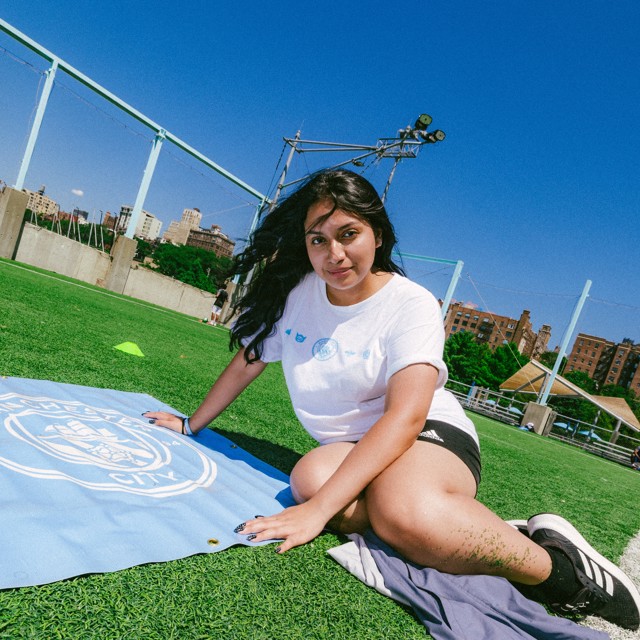 A woman sitting on a grassy field with a New York City flag in front of her, wearing a white shirt and shorts. The scene is outdoors on a sunny day.