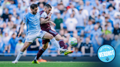 Two soccer players contesting a ball during a match, one in a light blue jersey and the other in maroon. Crowded stadium in the background with an 'Official Verdict' logo.